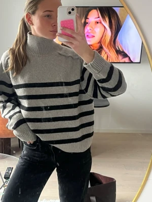 Grå stickad polotröja med svarta ränder - Säljer en oversized grå stickad polotröja med svarta horisontella ränder. Tröjan har breda ärmar, ribbade muddar och hög krage. Perfekt för dig som gillar en avslappnad och trendig look. Materialet känns mjukt och skönt mot huden.