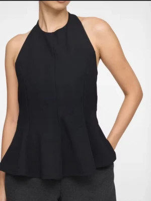 Svart ärmlös halterneck topp - Snygg svart halterneck topp med utsvängd nederdel och öppen rygg. Toppen är ärmlös och har en elegant, figurnära passform som ger en stilren siluett. 