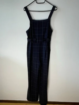 Mörkblå rutig jumpsuit med vida ben - Snygg mörkblå jumpsuit med vita och röda rutmönster. Har breda axelband, raka vida ben och markerad midja med bältesdetaljer. Materialet känns mjukt och följsamt.