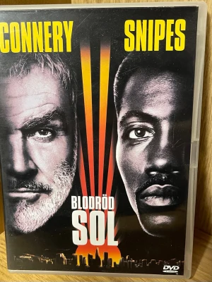 Blodröd sol DVD thriller - DVD-film med titeln 'Blodröd sol', baserad på Michael Crichtons bok. Omslaget har starka färger i svart, rött och gult med stadssilhuett och solstrålar. Filmen är en thriller med bonusmaterial som intervjuer och biotrailer. Svensk text och tal finns.