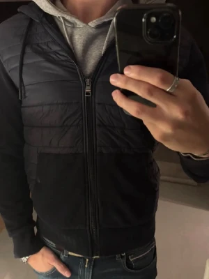 Svart cardigan från Tommy Hilfiger - Svart pufferjacka från Tommy Hilfiger med quiltad framsida och ärmar i mjukt material. Jackan har huva, dragkedja och diskret logotyp på ärmen. Perfekt för dig som vill ha en stilren och bekväm jacka med snygga detaljer.