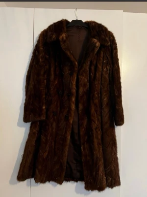 Mörkbrun vintage pälskappa - Snygg mörkbrun kappa i fuskpäls med bred krage och dubbelknäppning. Lång modell som ger en lyxig och retro känsla. Perfekt för dig som vill sticka ut och hålla dig varm under kalla dagar. Fodrad insida för extra komfort.