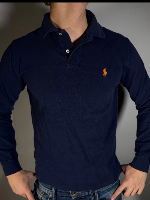 Marinblå långärmad skjorta Polo Ralph Lauren - Klassisk marinblå långärmad skjorta från Polo Ralph Lauren med orange broderad logga på bröstet. Skjortan har krage, knappar framtill och är tillverkad i mjuk bomull. Perfekt för en stilren och avslappnad look.