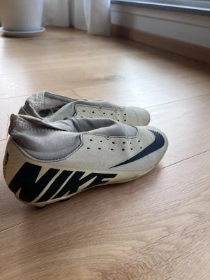Beige Nike Mercurial fotbollsskor - Säljer ett par beige Nike Mercurial fotbollsskor med svart logga och sula med fasta dobbar. Skorna har en strumpliknande passform och är tillverkade i syntetmaterial för bra grepp och kontroll på planen. Perfekta för dig som vill ha stilrena och funktionella skor.