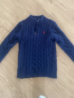Mörkblå kabelstickad tröja Polo Ralph Lauren - Kabelstickad mörkblå tröja från Polo Ralph Lauren med halv dragkedja och röd broderad logga på bröstet. Tröjan har ribbade muddar och hög krage, perfekt för kyliga dagar. Klassisk och tidlös design som passar till det mesta. Är XL i barnstorlek men skulle säga att den passar till ungefär mellan 173-180cm. Ungefärlig storlek herr XS-S