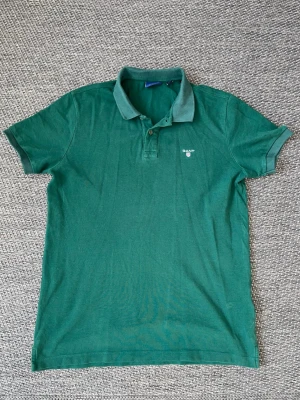 Grön pikétröja från Gant - Klassisk grön pikétröja från Gant med korta ärmar och krage. Tröjan har knappar vid halsen och en liten broderad logga på bröstet. Perfekt till jeans eller shorts för en avslappnad stil.