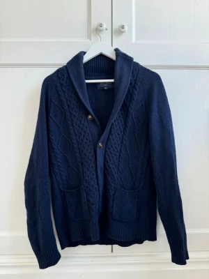 Mörkblå stickad kofta med sjalkrage - Mysig mörkblå stickad kofta med sjalkrage och två framfickor. Koftan har ett snyggt kabelstickat mönster framtill och stängs med knappar. Perfekt för lager på lager och ger en avslappnad men stilren look.