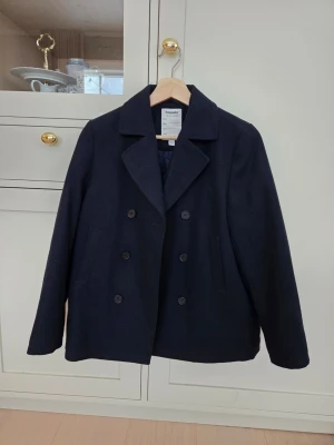 Marinblå skepparkavaj stl 152, kille - Stilren marinblå skepparkavaj från Kappahl med dubbelknäppning och bred krage. Klassisk modell med två sidofickor och slät yta. Perfekt för den som gillar tidlös och enkel stil. Gjord i ett mjukt material som känns bekvämt och håller värmen. Nyskick och säljes pga fel storlek. 