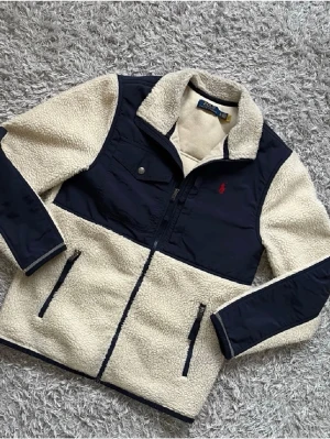 Beige fleece jacka från Polo Ralph Lauren - Säljer en beige fleece jacka från Polo Ralph Lauren med marinblå detaljer på bröst, ärmar och axlar. Jackan har dragkedja framtill, bröstficka med knapp, två sidofickor med dragkedja och en liten röd logga på bröstet. Ska även tilläggas att jackan passar både XS/S. Jackan är även Perfekt för en kall mysig vinterdag eller bara en mysig dag på hösten. Skriv vid funderingar😊