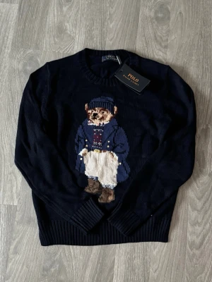 Mörkblå stickad tröja Polo Bear - Mörkblå stickad tröja från Polo Ralph Lauren med den ikoniska Polo Bear på bröstet. Tröjan har rund halsringning, ribbade muddar och är tillverkad i mjukt material. Perfekt för dig som gillar klassisk och lekfull stil.