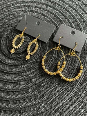 Två par guldfärgade örhängen med pärlor - Säljer två par örhängen i guldfärg. Ena paret är mindre hoops med små guldfärgade pärlor och en gnistrande sten längst ner. Det andra paret är större hoops med guldfärgade pärlor runt hela ringen. Perfekta för dig som gillar detaljerade accessoarer.