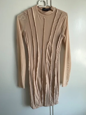 Beige långärmad klänning PrettyLittleThing - Trendig beige kortklänning från PrettyLittleThing med långa ärmar och coola vertikala sömdetaljer. Tillverkad i ett stretchigt syntetmaterial som sitter snyggt på kroppen. Perfekt för dig som gillar minimalistisk och modern stil.