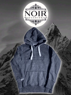 Mörkgrå hoodie från Superdry - Mörkgrå hoodie med huva och vita dragsnören från Superdry. Klassisk känguruficka framtill och ribbade muddar vid ärmslut och nederkant. Tillverkad i mjukt sweatshirtmaterial som är perfekt för en avslappnad stil.