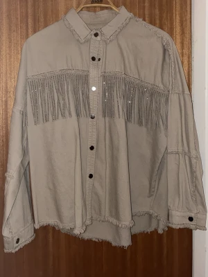 Beige skjorta med fransar och nitar - Cool beige skjorta med fransar och små nitar över bröstet. Skjortan har tryckknappar framtill, råa kanter och avslappnad passform. Tillverkad i ett kraftigare bomullstyg som ger en snygg oversized look. Perfekt för dig som gillar western-inspirerad stil.