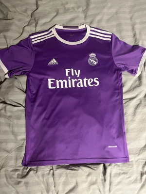 Lila Real Madrid matchtröja Adidas - Snygg lila Real Madrid matchtröja från Adidas med vita detaljer och klassiska ränder på axlarna. Tröjan har klubbmärke, Adidas-logga och Fly Emirates-tryck på bröstet. Tillverkad i lätt och ventilerande climacool-material, perfekt för fotbollsträning eller match.