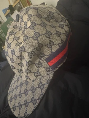 Gucci keps med monogram och röd-blå rand - Säljer en beige keps från Gucci med klassiskt blått GG-monogram över hela kepsen och en ikonisk röd-blå rand på sidan. Kepsen är tillverkad i canvasmaterial och har en mörkblå insida. Perfekt för dig som vill ha en lyxig och trendig accessoar.
