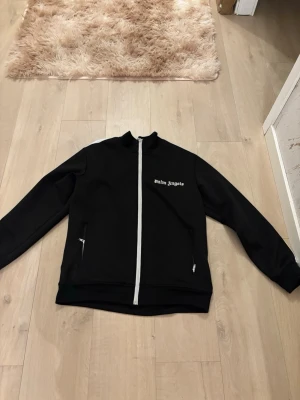 Svart zip hoodie från Palm Angels - Snygg svart hoodie från Palm Angels med vit dragkedja och vita detaljer på axlarna. Klassisk logga på bröstet och två fickor med dragkedja framtill. Tillverkad i ett mjukt och bekvämt material, perfekt för en streetwear-look.
