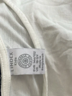 Vit topp med pärlor och strass Lindex - Söt vit topp från Lindex dekorerad med pärlor och strass i ett snirkligt mönster på framsidan. Tillverkad i mjuk bomull som känns skön mot huden. Perfekt för dig som vill ha något extra till din outfit.