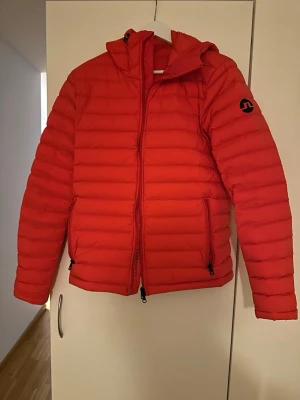Röd dunjacka från J.Lindeberg - Snygg röd dunjacka från J.Lindeberg med huva och svarta detaljer. Jackan har quiltad design, två sidofickor med dragkedja och logga på ärmen. Perfekt för kalla dagar och riktigt lätt att bära. Klassisk passform och modern look. Jacka är vind och vattentät Storlek M