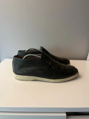 Svarta loafers i läder - Klassiska svarta loafers i äkta läder med slät ovandel och diskreta sömmar. Skorna har en låg vit sula och enkel slip-on design som gör dem lätta att ta på. Perfekta för en stilren och avslappnad look.