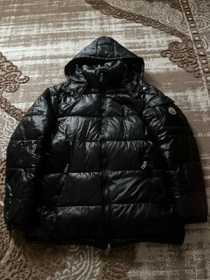 Svart dunjacka från Moncler med huva - Snygg svart dunjacka från Moncler med glansig finish och huva. Jackan har två dragkedjeförsedda fickor framtill och Moncler-logga på ärmen. Perfekt för kalla vinterdagar och riktigt varm tack vare dunfyllningen.