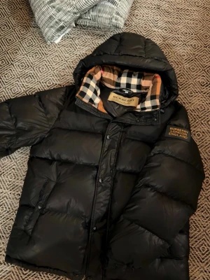 Svart pufferjacka från Burberry - Säljer en svart pufferjacka från Burberry med klassiskt rutigt foder i beige, svart och rött. Jackan har huva, dragkedja och tryckknappar samt fickor framtill. Märkeslogga på ärmen och oversized look. Perfekt för kalla vinterdagar.