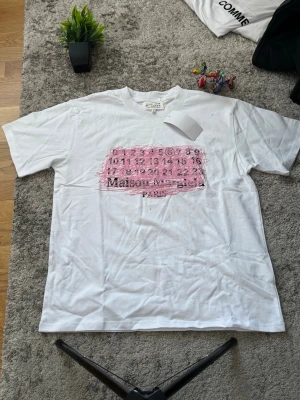  Maison Margiela t-shirt - Vit t-shirt från Maison Margiela med ett stort rosa tryck framtill som visar märkets ikoniska siffror och logga. Klassisk passform och rund hals. Tillverkad i mjuk bomull, perfekt för en clean och trendig look.