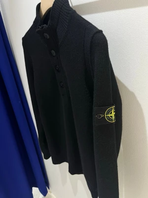 Svart stickad tröja Stone Island - Svart stickad tröja från Stone Island med hög krage och fyra knappar framtill. Klassisk patch med logga på vänster ärm. Tröjan har lång ärm och ribbade muddar vid hals och ärmslut. Perfekt för dig som gillar streetwear och stilren design. Endast använt 1 gång.