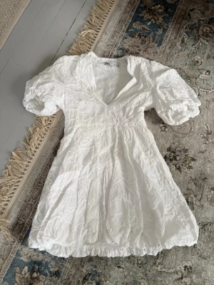 Vit kort klänning från Zara - Superfin vit kort klänning från Zara med puffärmar och v-ringning. Klänningen har broderade blommönster och en lätt volang nertill. Perfekt för studenten. Finns även dragkedja på sidan (se bild). Ingen prislapp kvar men är aldrig använd, köpt i somras.
