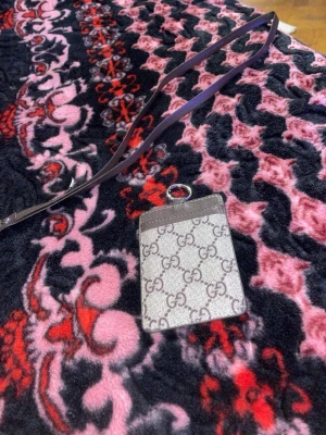 Gucci kortfodral med halsband - Snyggt kortfodral från Gucci med klassiskt GG-mönster i grått och beige. Fodralet har ett fack för kort och ett långt brunt läderband som kan bäras runt halsen. Perfekt för att hålla dina kort nära till hands och samtidigt visa upp en trendig accessoar.