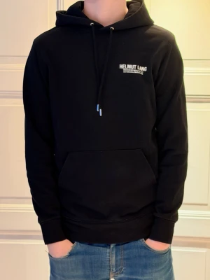Svart hoodie från Helmut Lang - (Moddelen är 185-186 och väger 75 kg)Snygg svart hoodie från Helmut Lang med vit logga på bröstet. Klassisk känguruficka framtill, justerbar dragsko i huvan och ribbade muddar vid ärmar och midja. Tillverkad i mjukt material som känns skönt mot huden. Perfekt för en avslappnad och trendig look.