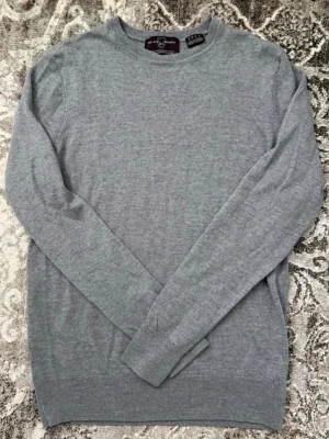 Grå merinoulltröja från Black Brown - Säljer en stilren grå stickad tröja från Black Brown 1826. Tröjan är tillverkad i 100% extrafin merinoull, har rund halsringning och långa ärmar. Perfekt för dig som gillar klassiska och mjuka plagg.