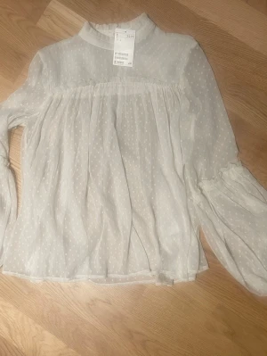 Vit transparent blus från H&M stl 34 - Söt vit blus från H&M i transparent, tunn och luftig polyester med diskret prickmönster. Blusen har hög krage, långa puffärmar med resår vid ärmslut och en romantisk, flowy passform. Perfekt för att skapa en trendig och feminin look.