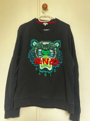 Svart Kenzo sweatshirt med tiger - Svart sweatshirt från Kenzo med ikoniskt broderat tigerhuvud i grönt, blått och vitt samt röd Kenzo-logga på bröstet. Rund halsringning och ribbade muddar. Perfekt statement-plagg för dig som gillar streetwear.