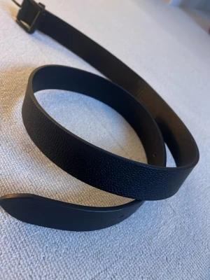 Svart klassiskt skärp i skinn(. Helt oanvänd). - Snyggt svart bälte i äkta skinn med enkel, matt svart metallspänne. Stilren design som passar till jeans eller kostymbyxor. Bältet har en lätt texturerad yta och är smalt, vilket gör det lätt att matcha med olika outfits.