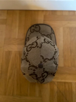 Beige GG-mönstrad keps från Gucci - Säljer en beige keps från Gucci med det klassiska GG-monogrammet i brunt över hela kepsen. Kepsen har böjd skärm och justerbar baksida. Tillverkad i slitstarkt tyg med mörkbruna detaljer längs kanterna. Perfekt för dig som gillar exklusiva accessoarer.
