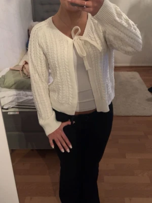 Vit kabelstickad kofta med rosett - Söt vit kabelstickad kofta med knytband i halsen och långa ärmar. Koftan har en klassisk stickad struktur och ribbade muddar vid ärmslut och nederkant. Perfekt att slänga över en topp för en chill look.