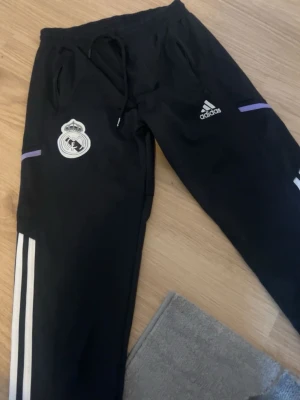 Svarta Real Madrid träningsbyxor Adidas - Säljer ett par svarta träningsbyxor från Adidas med Real Madrid-logga på ena benet. Byxorna har vita ränder längs sidorna, lila detaljer och dragsko i midjan. Perfekta för fotbollsträning eller chill. Materialet är troligtvis polyester.