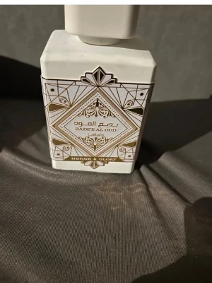 Badee Al Oud Honor & Glory parfym - Säljer en elegant parfym från Lattafa, Badee Al Oud Honor & Glory. Flaskan är vit med guldiga och svarta geometriska mönster och har en fyrkantig form. Designen ger en lyxig känsla och flaskan är tillverkad i plast eller glas med dekorativ etikett.