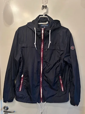 Marinblå vindjacka från Polo Ralph Lauren - Snygg marinblå vindjacka från Polo Ralph Lauren med huva och vita dragskor. Jackan har dragkedja framtill med röd och vit kant, två fickor med dragkedja och Polo-märke på ärmen. Tillverkad i lätt, vindtätt material som passar perfekt till blåsiga dagar.