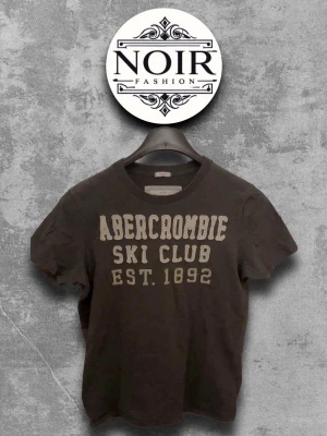 Svart Abercrombie t-shirt med tryck - Cool svart t-shirt från Abercrombie med beige texttryck 'SKI CLUB EST. 1892' på bröstet. Klassisk passform och rund halsringning, perfekt till jeans eller shorts. Skön bomullskvalitet som känns mjuk mot huden.