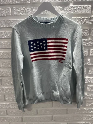 Ljusblå stickad tröja Ralph Lauren - Snygg ljusblå stickad tröja från Ralph Lauren med amerikansk flagga i rött, vitt och blått på bröstet samt RL-broderi. Tröjan har rund halsringning och ribbade muddar vid ärmslut och nederkant. Perfekt för dig som gillar klassisk preppy stil.