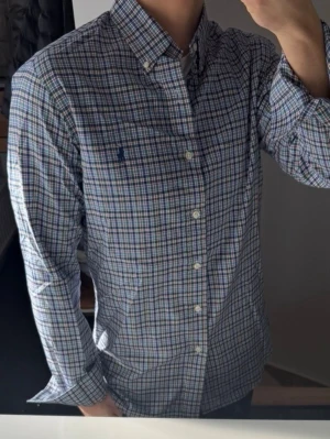 Rutig skjorta från Polo Ralph Lauren - Klassisk rutig skjorta från Polo Ralph Lauren i blå, grön och vit färg. Skjortan har lång ärm, knappar framtill och en liten broderad logga på bröstet. Tillverkad i mjuk bomull och har en normal passform. Perfekt för en stilren och avslappnad look.