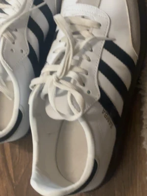 Adidas Samba vita sneakers - Klassiska Adidas Samba sneakers i vitt med svarta ränder och gulddetalj på sidan. Skorna har snörning och är tillverkade i skinn med mockadetaljer på tån. Perfekta för en sportig och stilren look.