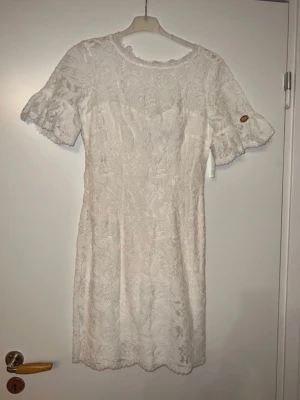 Vit spetsklänning/studentklänning från Chiara Forthi - Säljer en elegant vit kortklänning från Chiara Forthi med vackra spetsdetaljer och korta ärmar. Klänningen har rund halsringning och ett feminint, figurnära snitt. Perfekt för sommarens alla tillfällen!