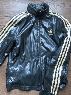 Träningsjacka - Klassisk svart vindjacka från Adidas med guldiga ränder längs ärmarna och Adidas-logga på bröstet 🖤