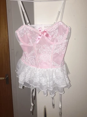 Rosa och vit spetskorsett med volang - Supersöt korsett i ljusrosa med vita spetsdetaljer och volangkant. Dekorerad med en liten rosa rosett framtill, justerbara axelband och strumpebandshållare. 