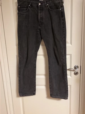 Svarta raka jeans med fem fickor - Säljer ett par svarta jeans med rak passform och klassisk femficksdesign. Jeansen har normal midja, bälteshällor och stängs med knapp och dragkedja. Materialet är denim i bomull och färgen är tvättad svart för en cool look.