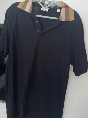 Svart pikétröja med rutig krage Burberry - Snygg svart pikétröja från Burberry med klassisk rutig krage i beige, rött och svart. Tröjan har korta ärmar och knäppning framtill med två knappar. Perfekt för en stilren och trendig look. Storlek M