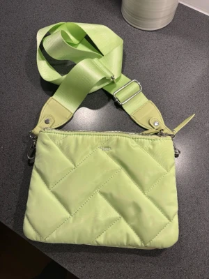 Ljusgrön quiltad axelväska från Don Donna - Trendig ljusgrön axelväska från Don Donna med quiltat mönster och bred justerbar axelrem i matchande färg. Väskan har silverfärgade metalldetaljer och dragkedja upptill. Perfekt för att addera färg till din outfit.